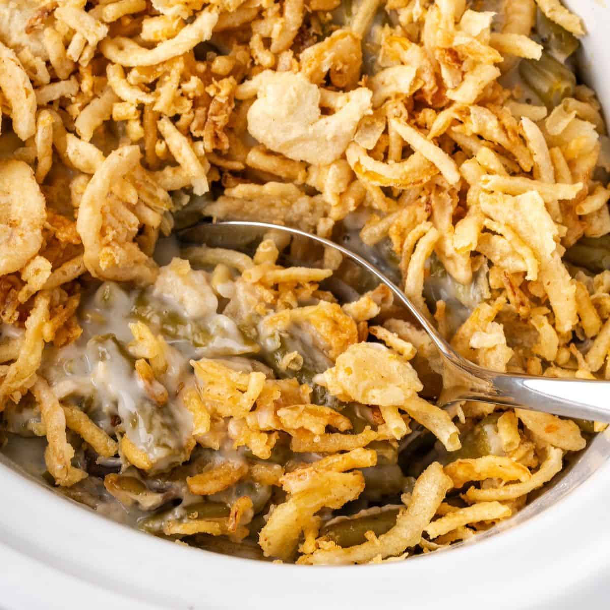 A silver serving spoon scooping up a serving of green bean casserole from a white crock pot.