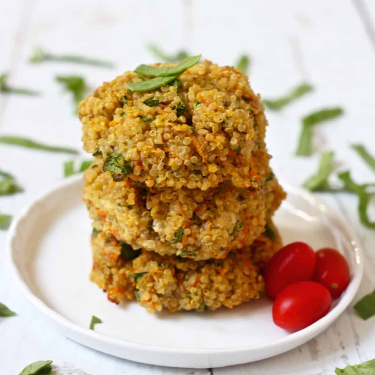 Close up of three veggie quinoa cakes stacked on a small white plate with cherry tomatoes to the side and basil sprinkled all around.