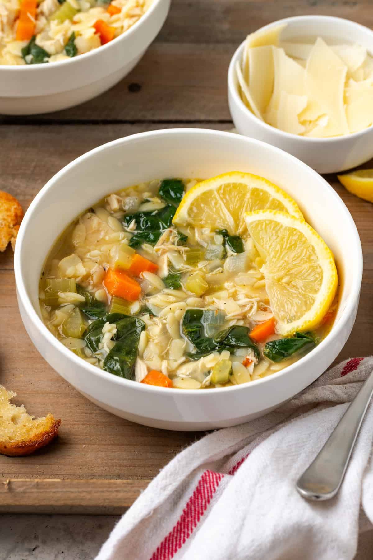 Lemony chicken orzo soup served in a white bowl with lemon slices to the side and a bowl of fresh shaved Parmesan in the background.