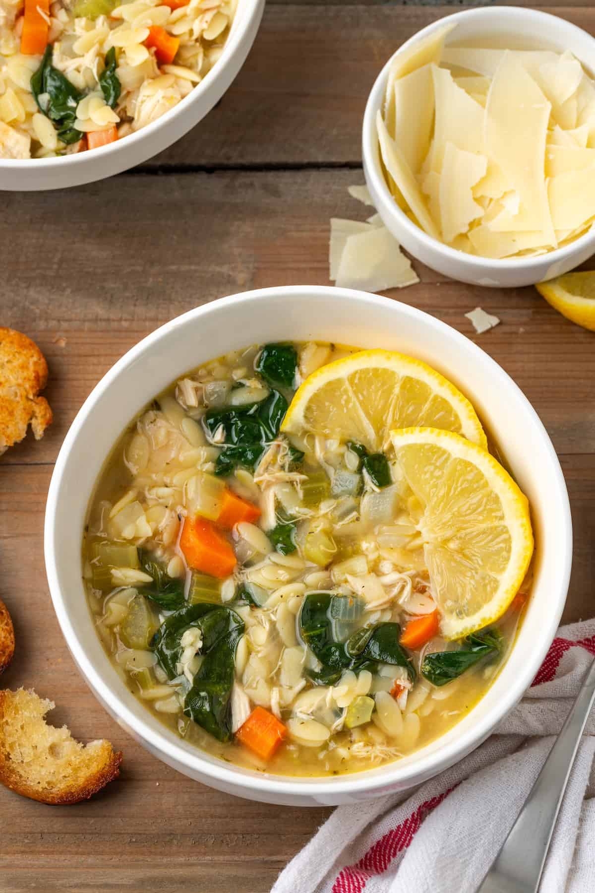 A bowl of lemon chicken orzo soup with spinach served in a white bowl with fresh shaved Parmesan and bread slices to the side.