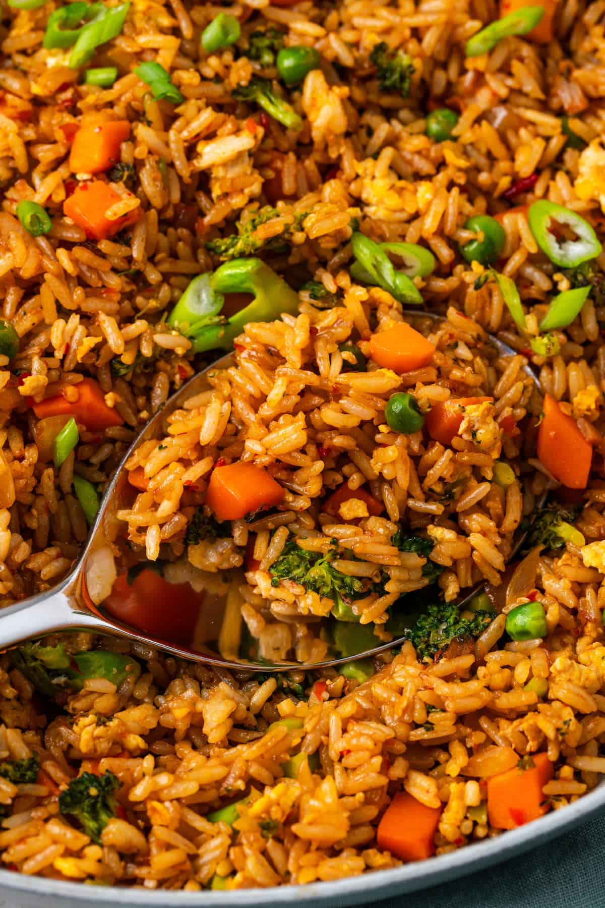 A spoon scooping up a serving of veggie fried rice from a large skillet.