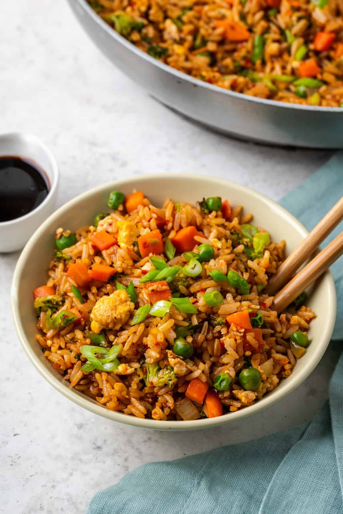 A beige bowl full of vegetable fried rice with chopsticks resting in it, a bowl of soy sauce to the side, and a skillet of fried rice in the background.