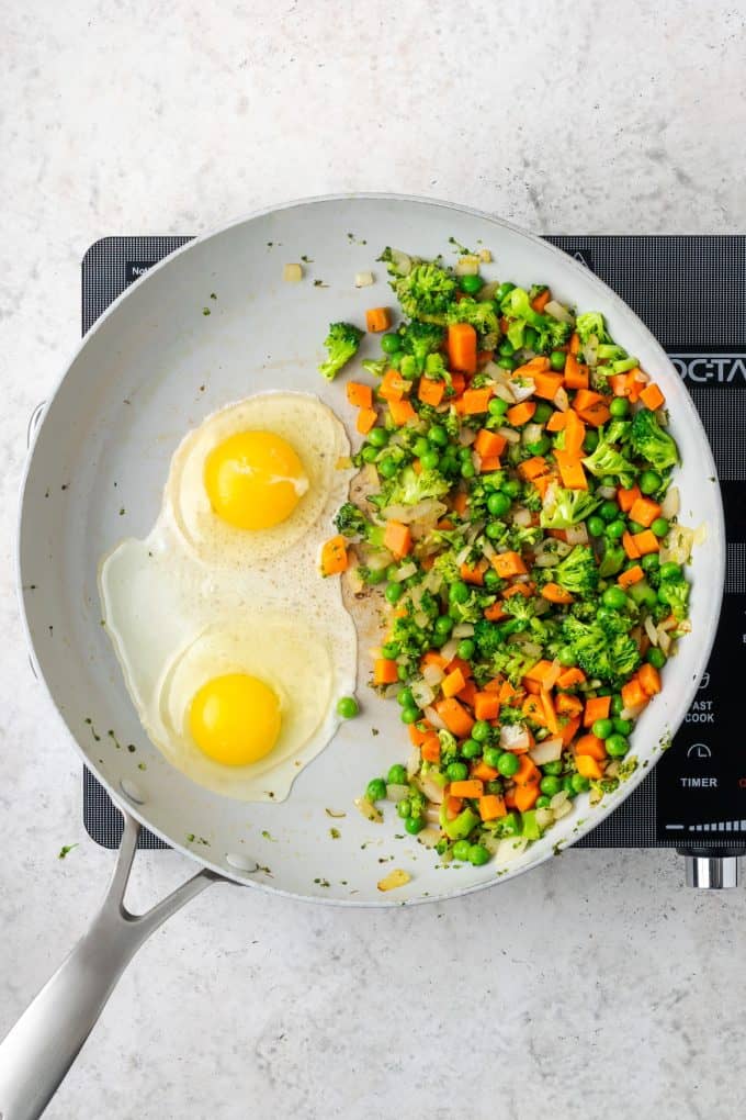 Sauteed veggies pushed to the side of a large skillet and two eggs cracked into the other side.