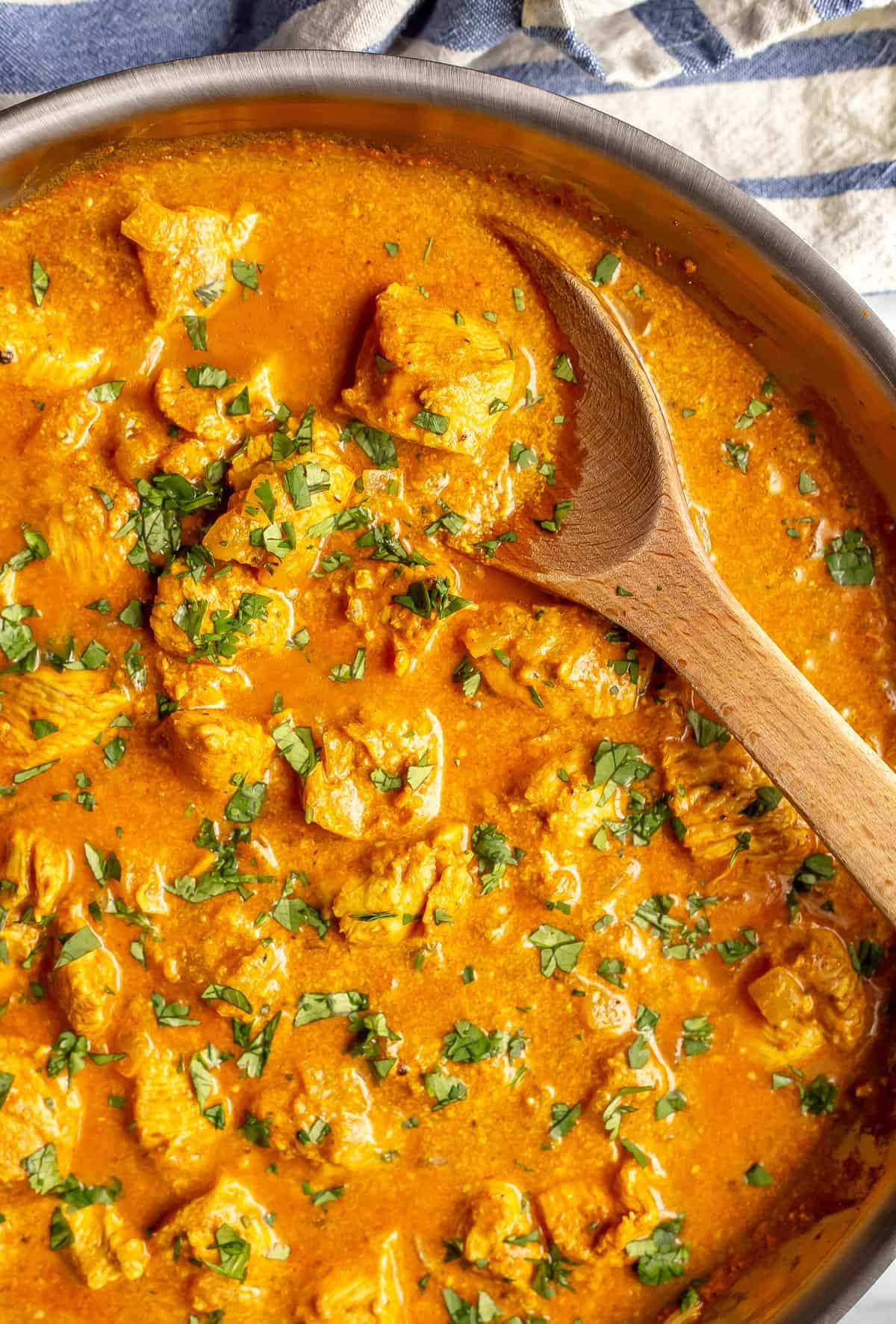 A wooden spoon resting in a large saute pan of butter chicken.