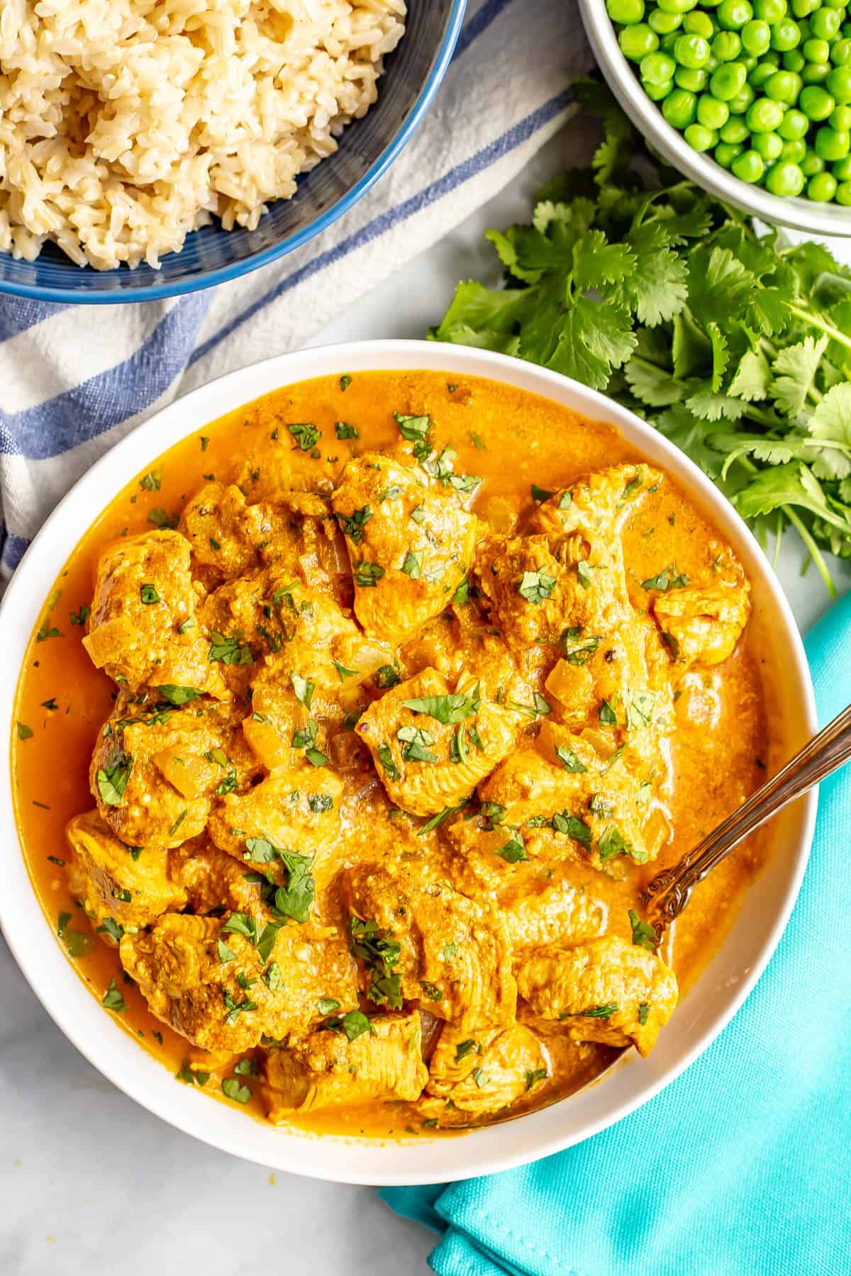 A spoon resting in a bowl of healthy butter chicken with bowls of rice and peas in the background.