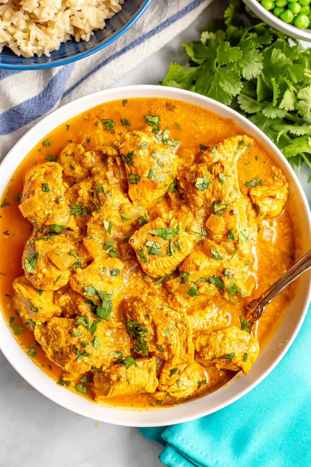 Close up of a spoon resting in a bowl of healthy butter chicken with bowls of rice and peas in the background.