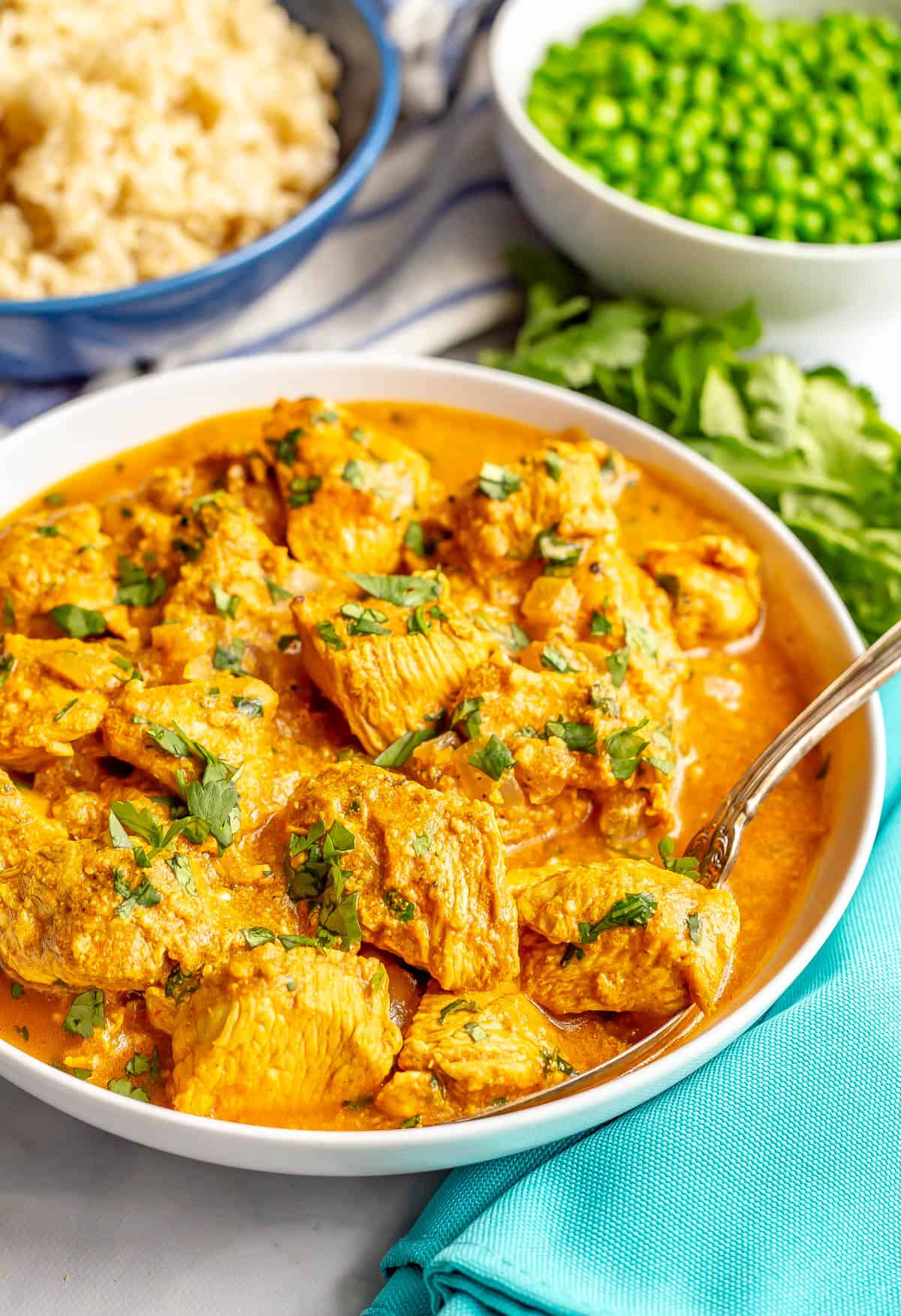 A serving spoon resting in a white bowl of Indian butter chicken with bowls of rice and peas in the background.