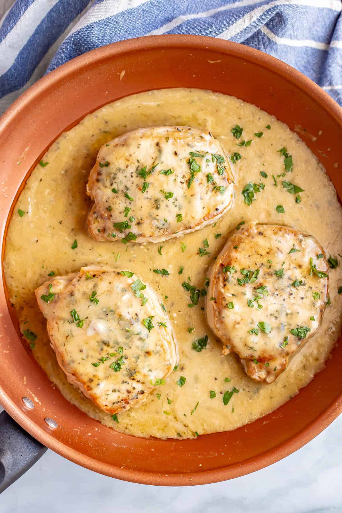 Three seared pork chops with gravy in a large skillet with a blue striped towel to the side.