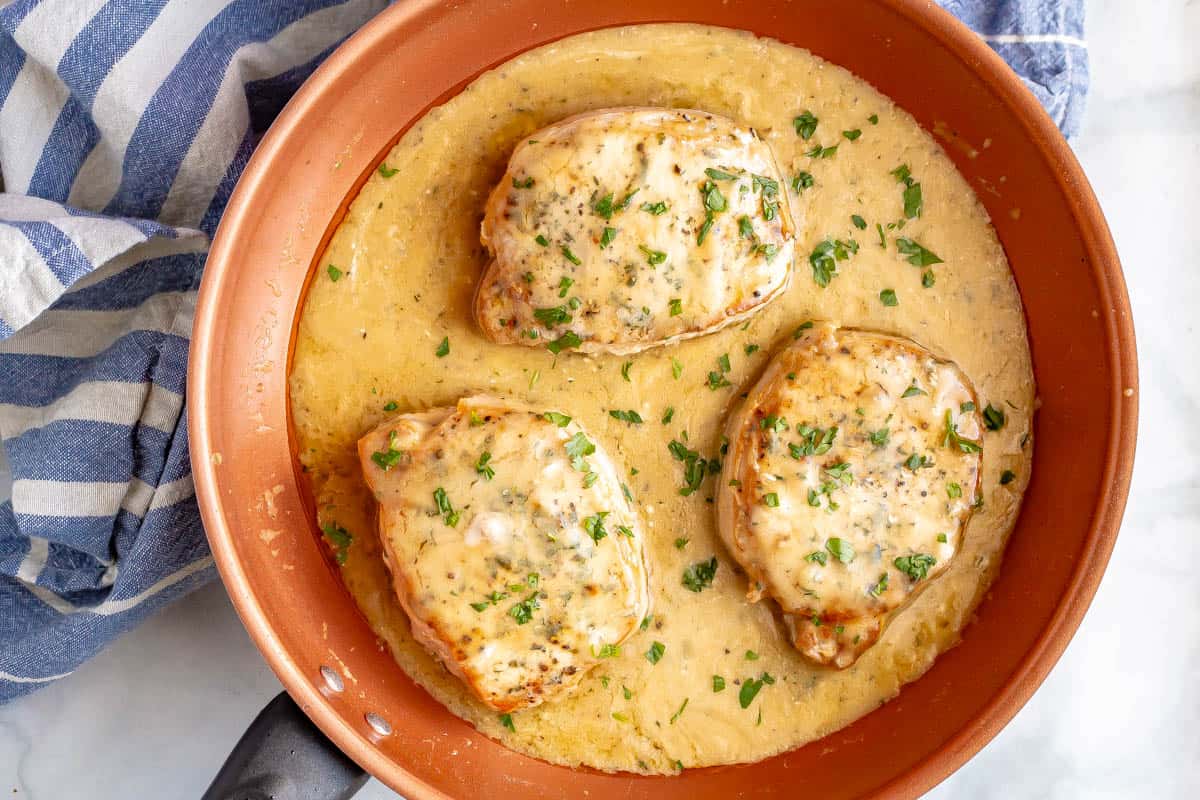 Three seared pork chops in a copper pan with gravy sprinkled with parsley.