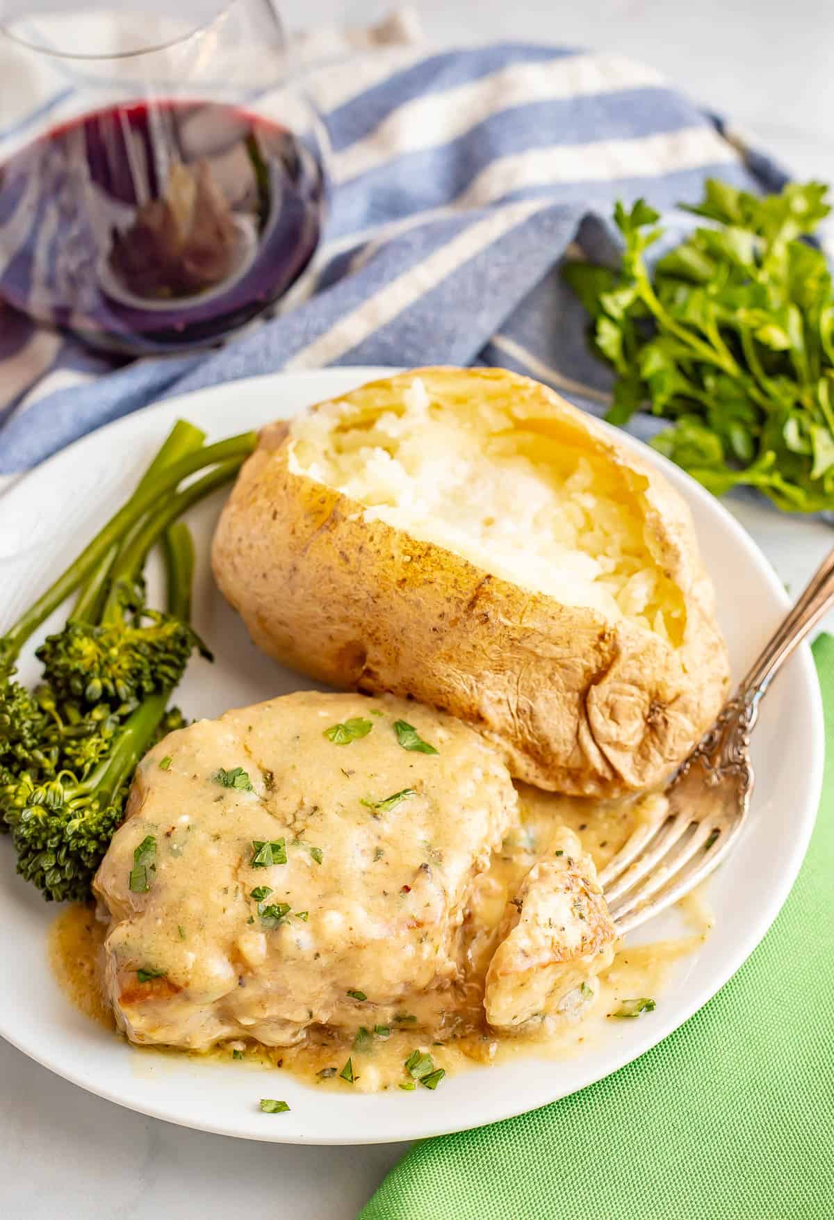 A white dinner plate with a baked potato, broccolini, pork chop with gravy and a fork resting on the side, with a glass of red wine in the background.