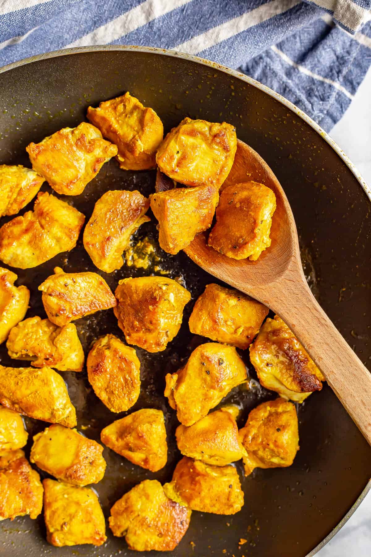 Cooked cubed turmeric chicken pieces scooped onto a wooden spoon resting in a large dark skillet.