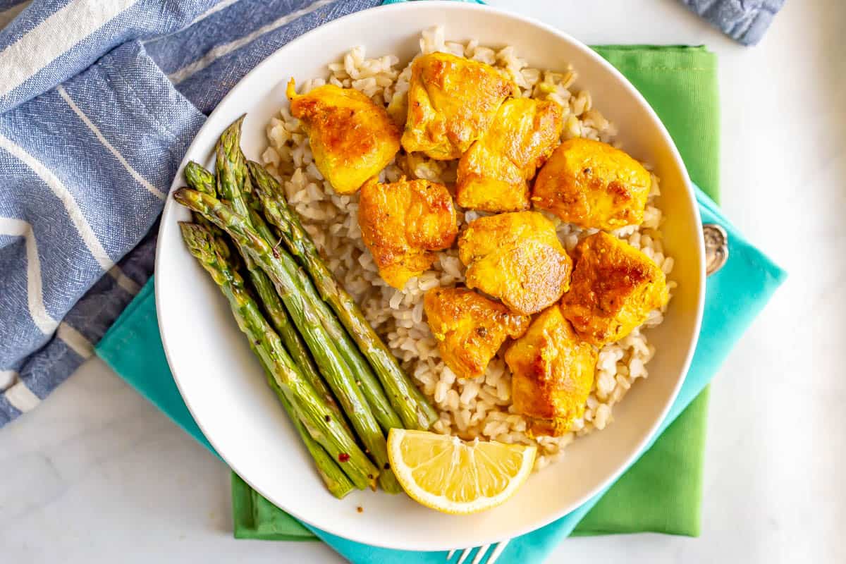 A low white bowl with rice topped with seasoned cubed chicken pieces and roasted asparagus on the side.