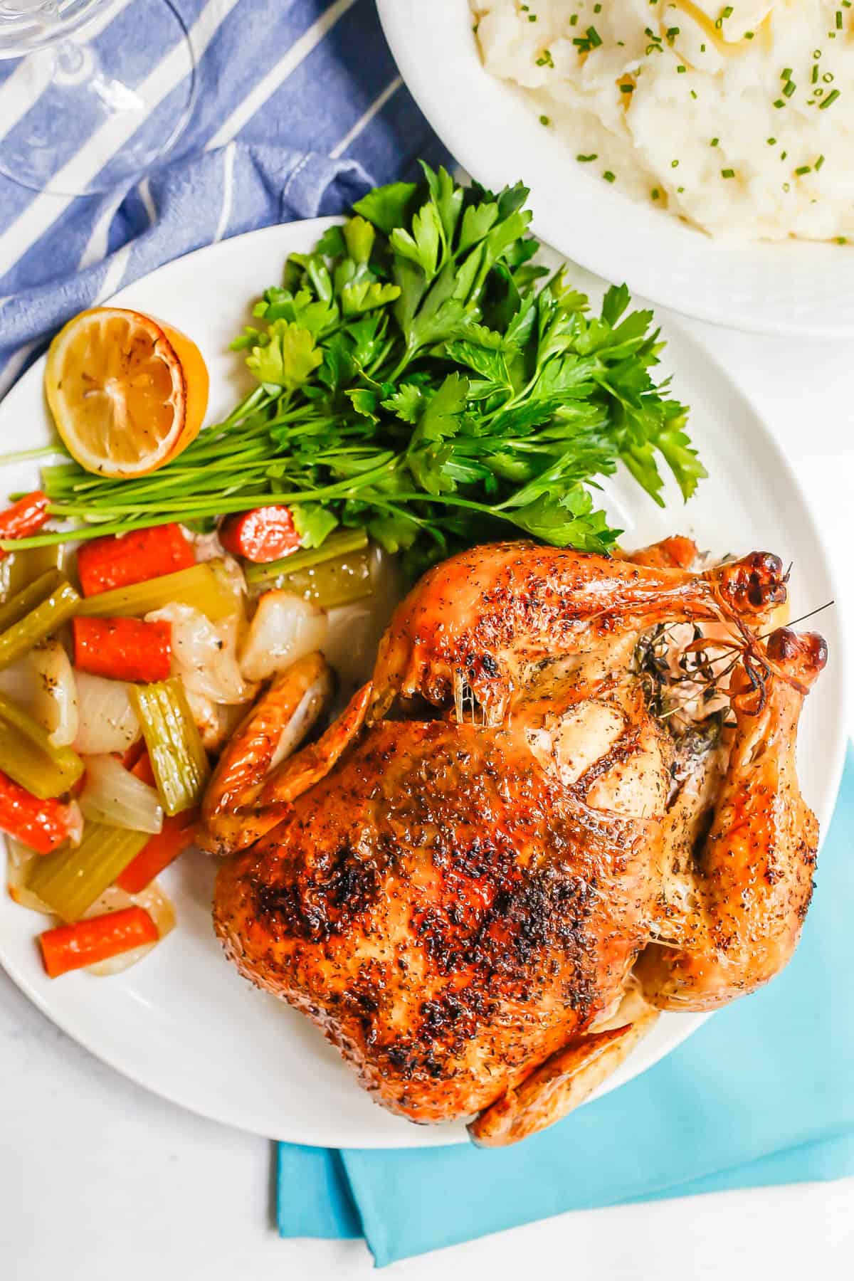 A whole roast chicken served on a large white platter with roasted veggies to the side and parsley for garnish.