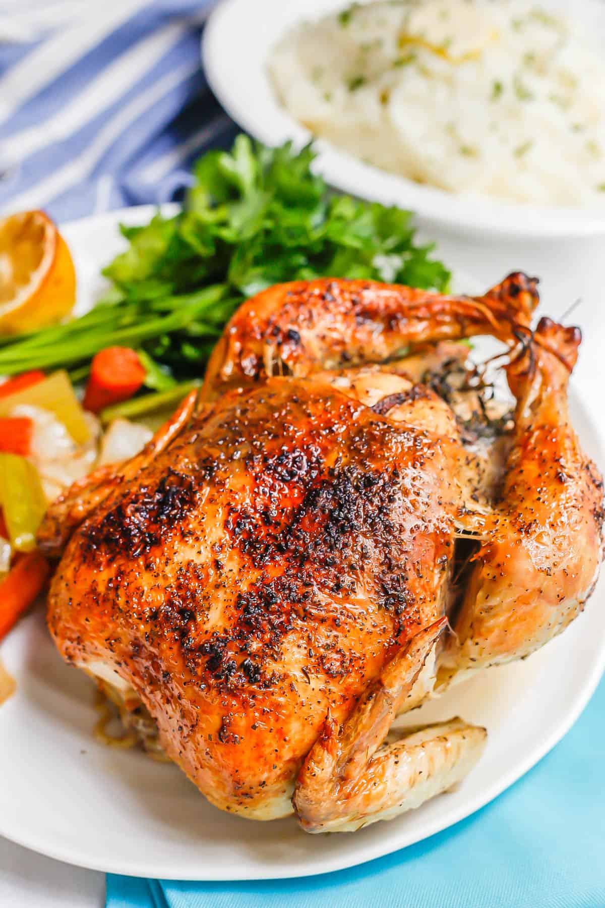 Close up of a browned, seasoned and oven roast chicken served on a white platter with a bowl of mashed potatoes in the background.
