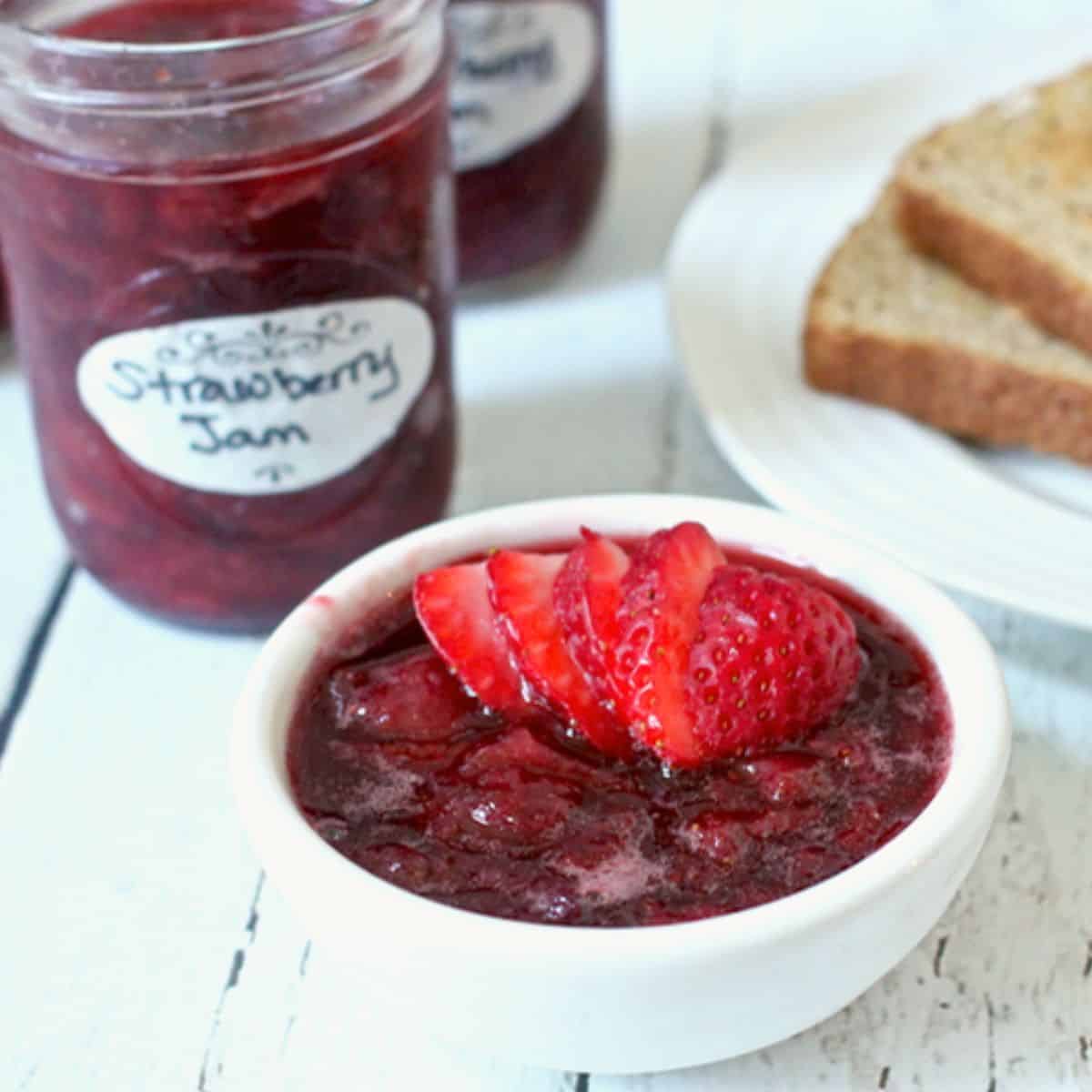 Homemade strawberry freezer jam served in a small white bowl with a jar of jam in the background beside a plate of toast.