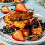 Powdered sugar being sprinkled onto French toast sticks arranged and stacked on a plate with fresh fruit scattered around and a text overlay on the photo.