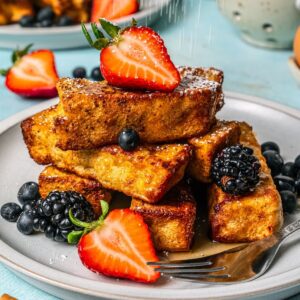 Close up of powdered sugar being sprinkled onto French toast sticks arranged and stacked on a plate with fresh fruit scattered around.