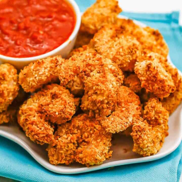 Close up of breaded shrimp piled on a white plate alongside a small bowl of marinara sauce for dipping.