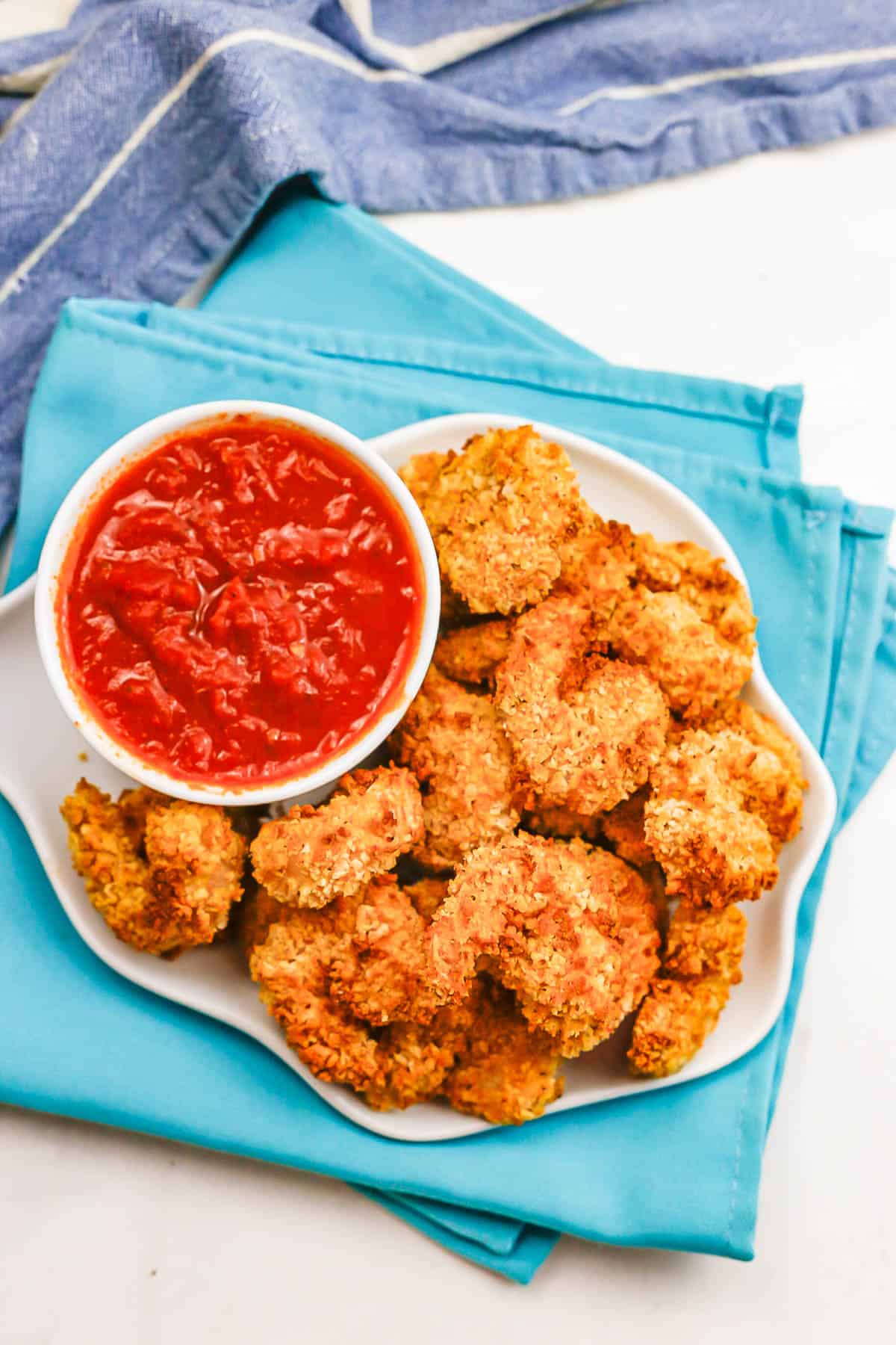 A white plate with breaded shrimp piled on beside a small bowl of marinara sauce for dipping.