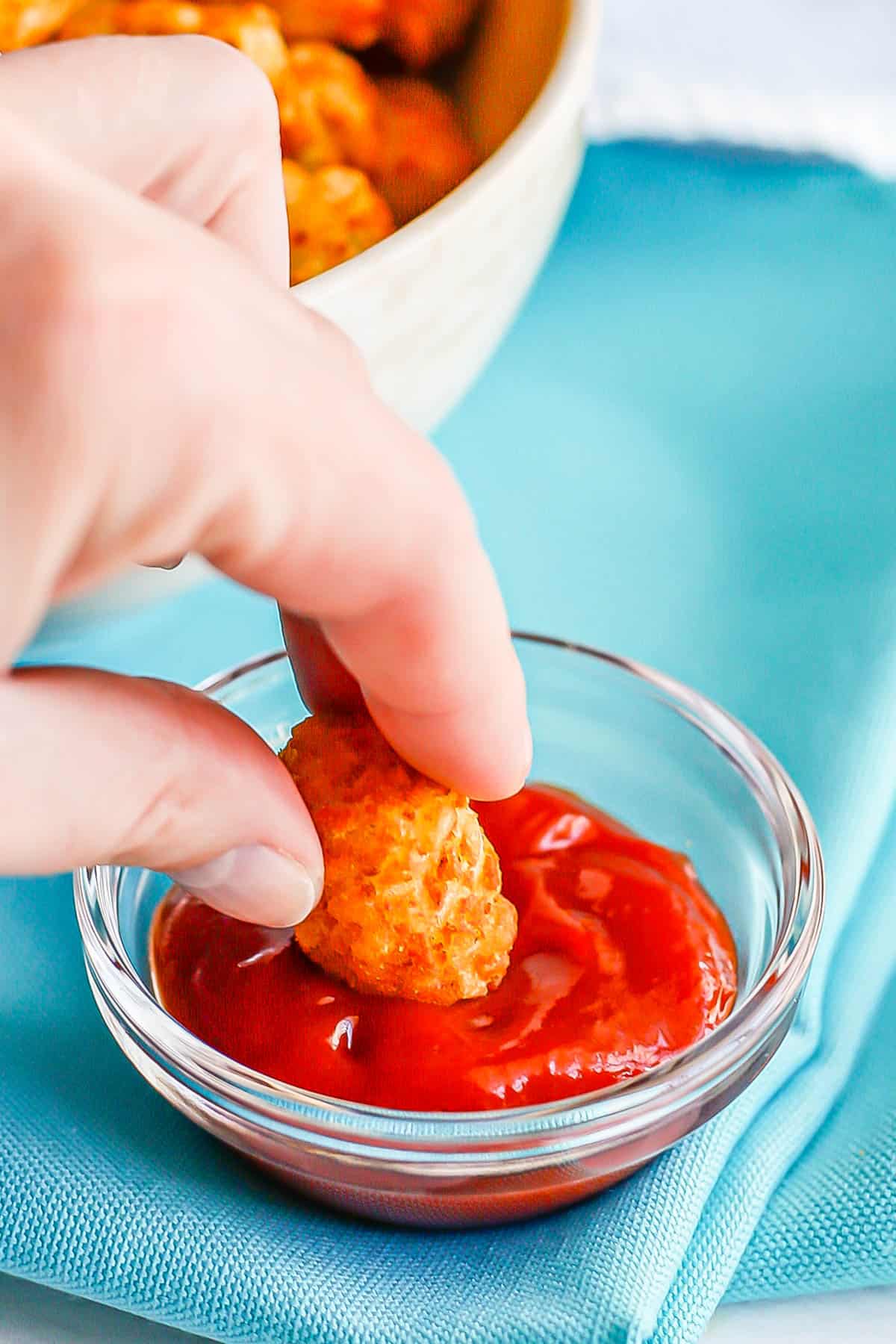 A hand dipping a tater tot into a small bowl of ketchup.