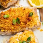 Close up of a crunchy buttery baked cod filet on a platter with sliced green onions on top and a lemon wedge to the side and a text overlay on the photo.