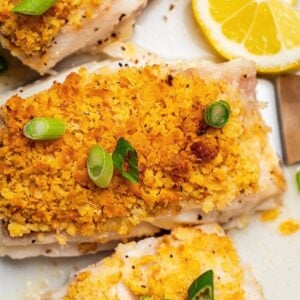 Close up of a crunchy buttery baked cod filet on a platter with sliced green onions on top and a lemon wedge to the side.