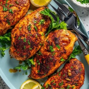 Close up of baked paprika chicken served on a teal platter with fresh parsley and lemon wedges and serving utensils to the side.