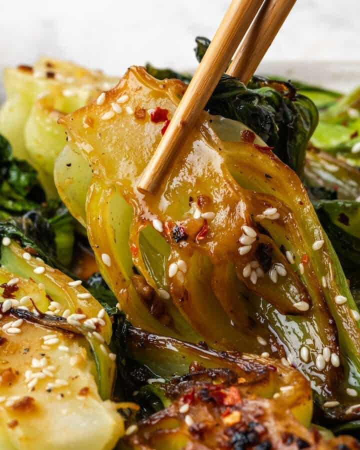 Close up of chopsticks lifting up a piece of baby bok choy with garlic and ginger from a serving platter.