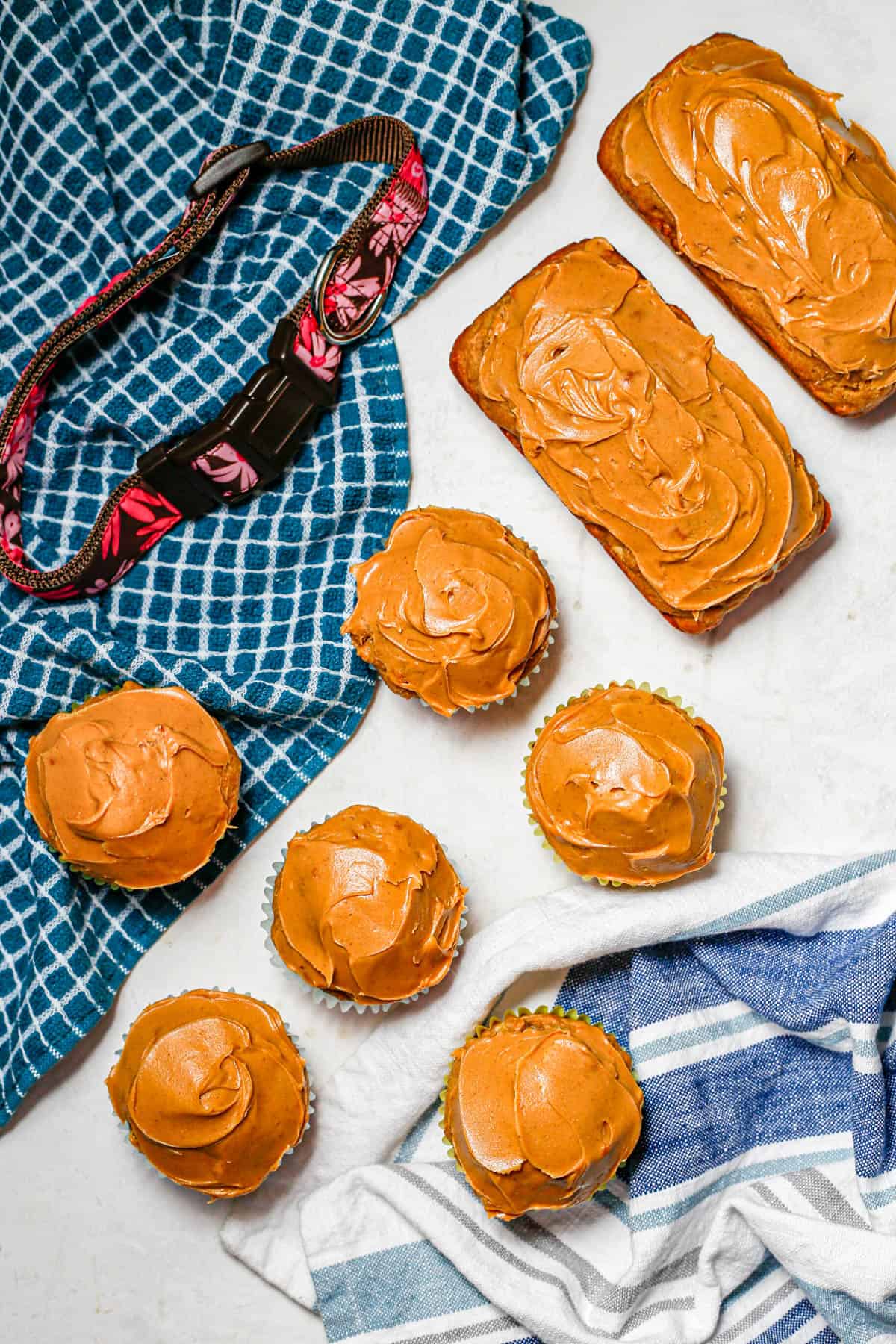Peanut butter frosted dog cupcakes and mini dog cakes on a counter beside kitchen towels and a dog collar.