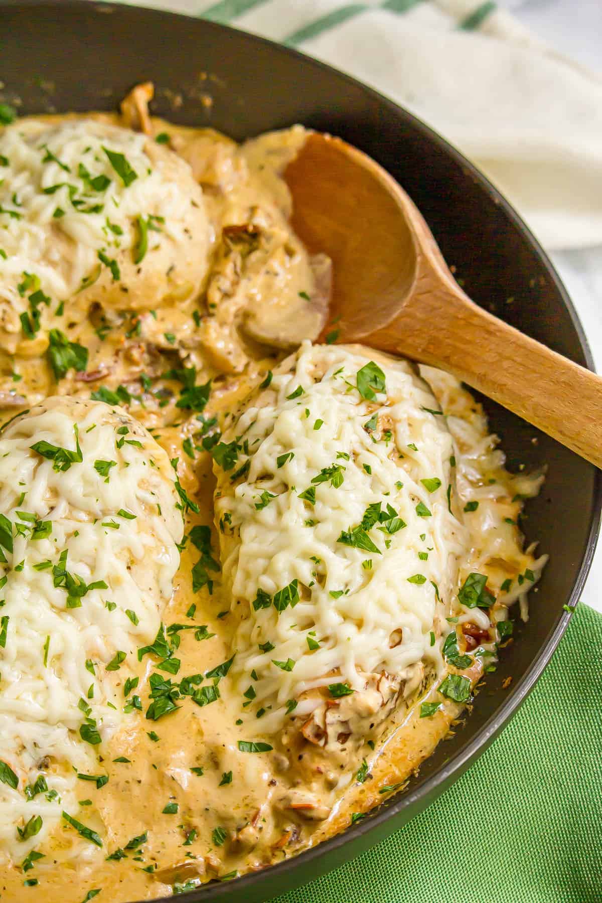 A wooden spoon resting in a large dark skillet with cheesy chicken breasts topped with chopped fresh parsley.