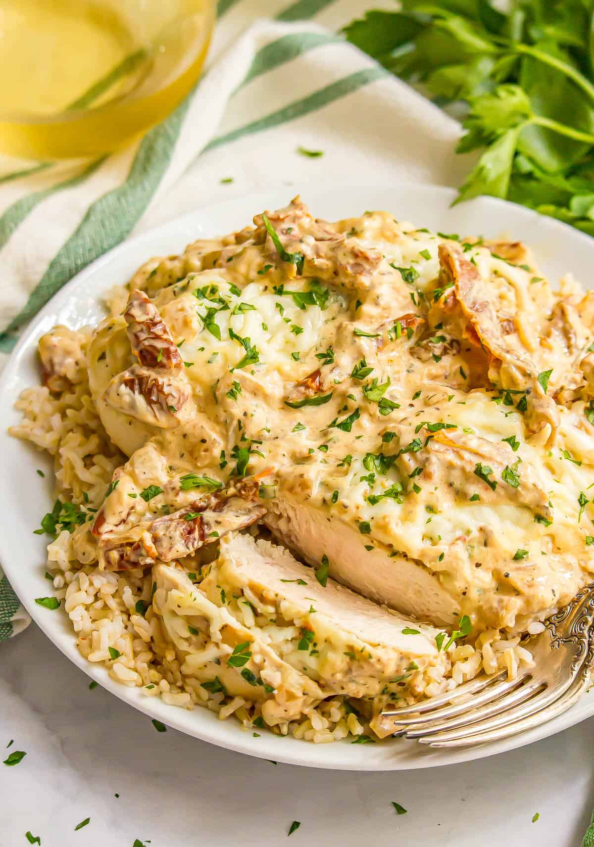 A fork resting on a white plate of a sliced creamy Italian chicken breast served over rice with parsley sprinkled on top.