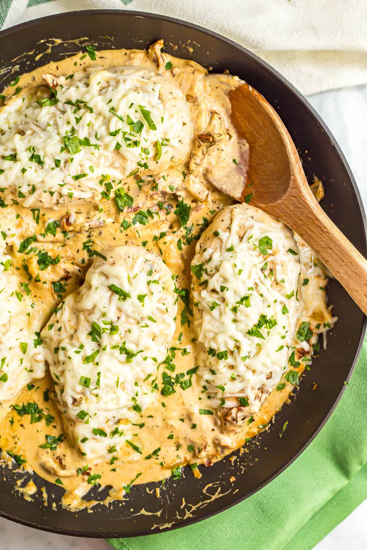 A wooden spoon resting in a large skillet with cheesy Italian chicken breasts in a creamy sauce.