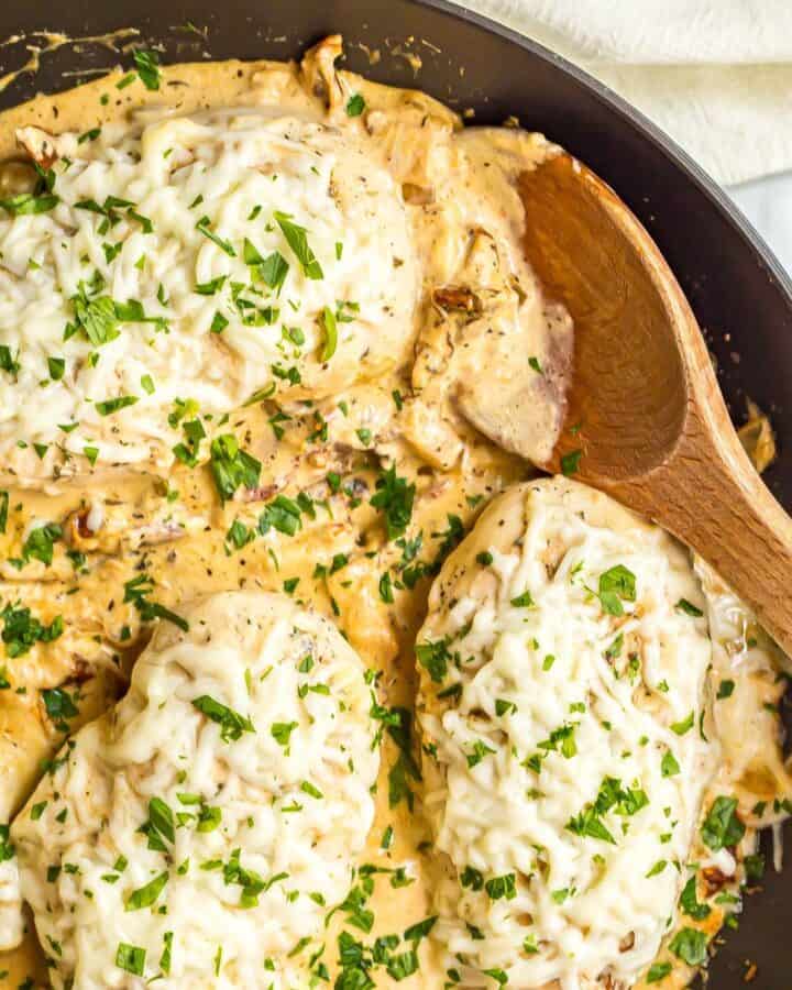Close up of a wooden spoon resting in a large skillet with cheesy Italian chicken breasts in a creamy sauce.