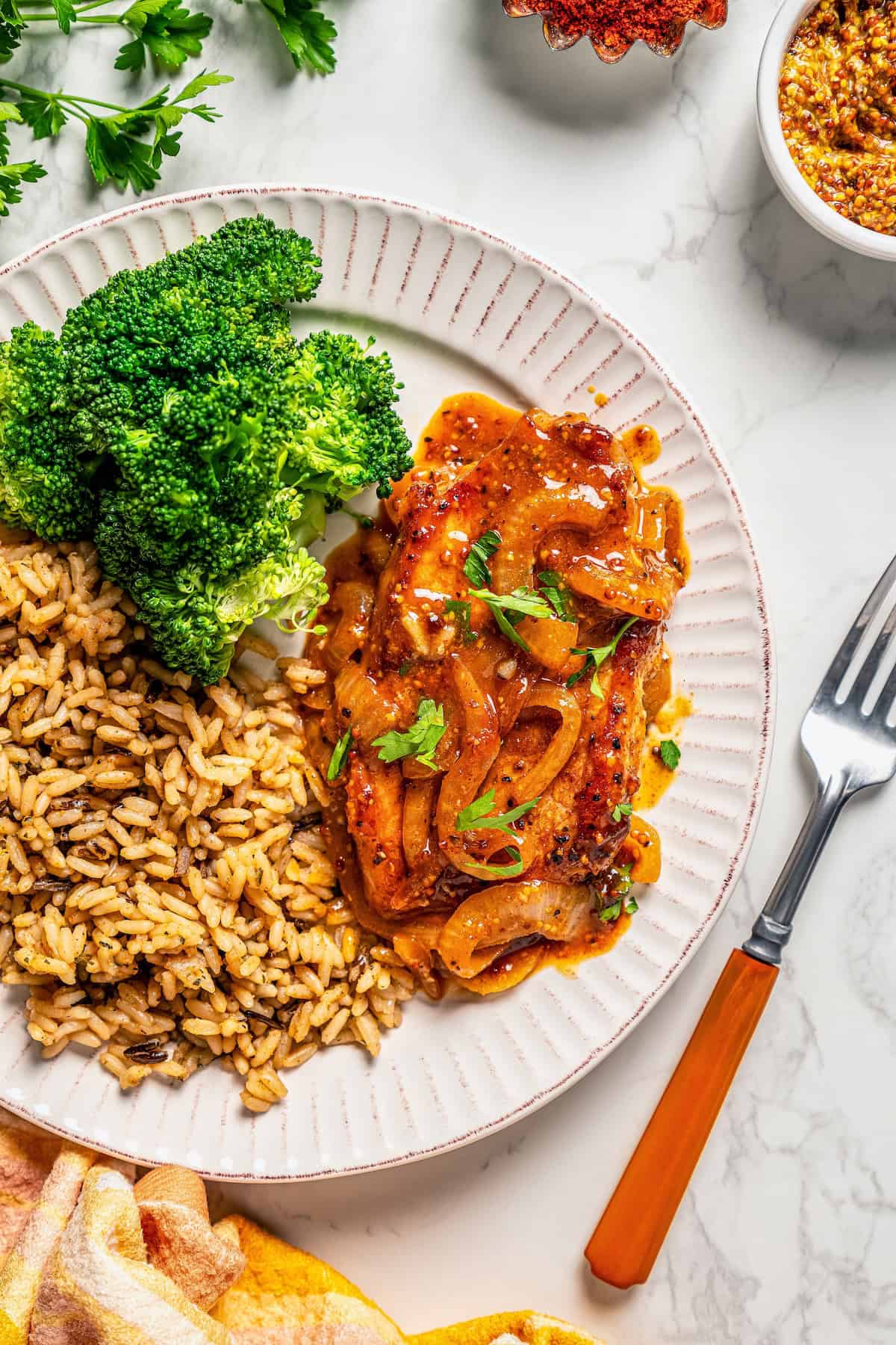 A pork chop with a honey mustard sauce and sautéed onions served with wild rice and broccoli on a plate.