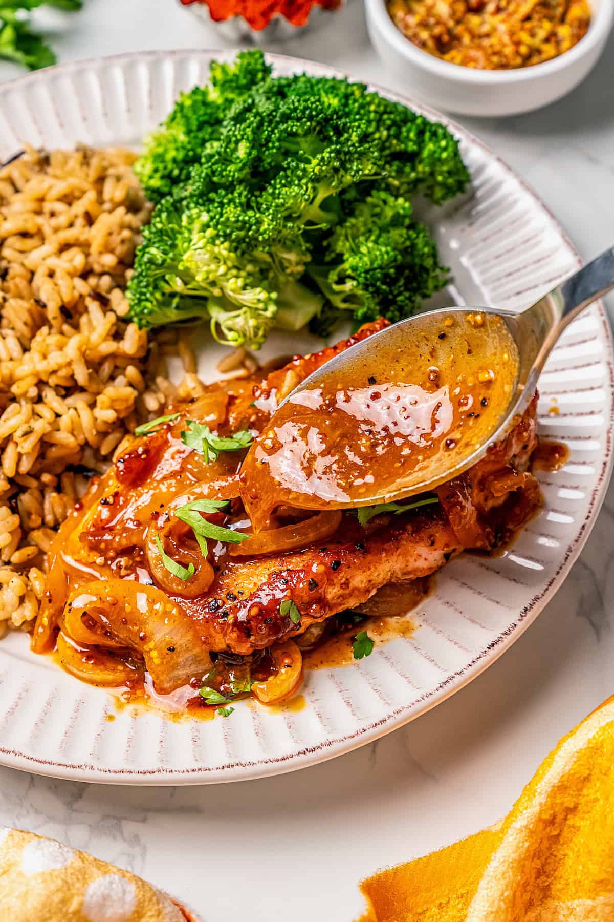 Sauce being spooned over a pork chop served with wild rice and broccoli on a plate.