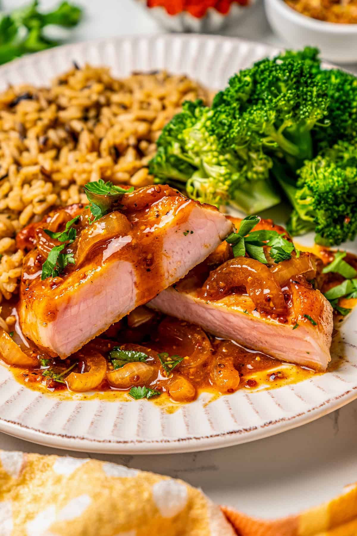 A pork chop cut in half and stacked on a dinner plate with sauce drizzled over top and rice and broccoli to the side.