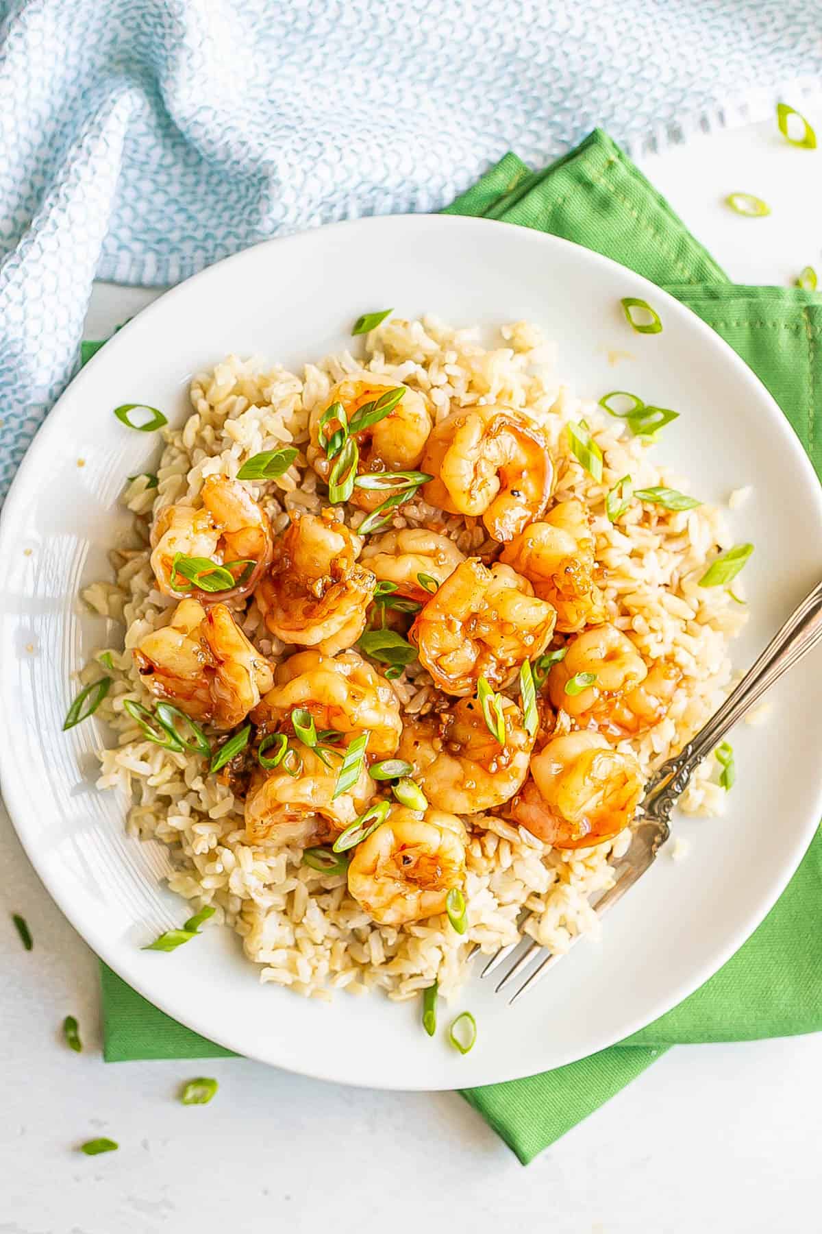A fork resting on a white round plate with rice topped with honey garlic shrimp and sprinkled with sliced green onions.