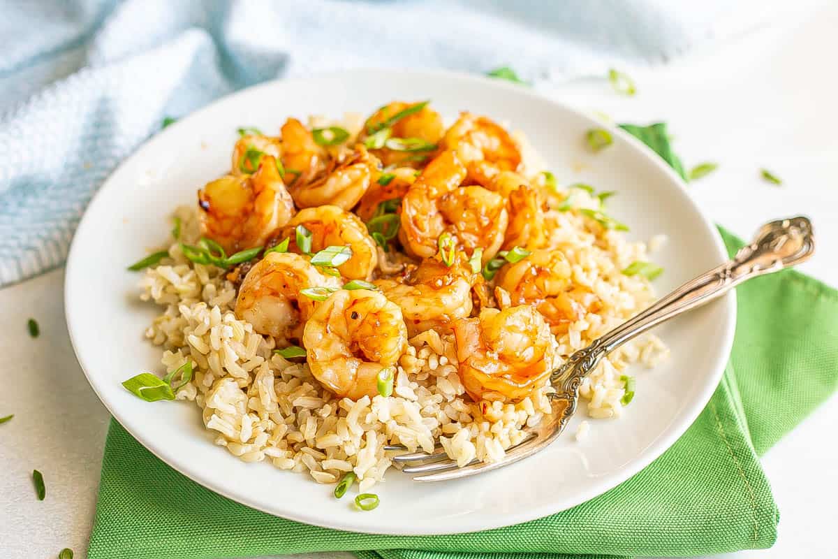 A fork resting on a white plate set on a green napkin with rice and shrimp in a honey garlic sauce.