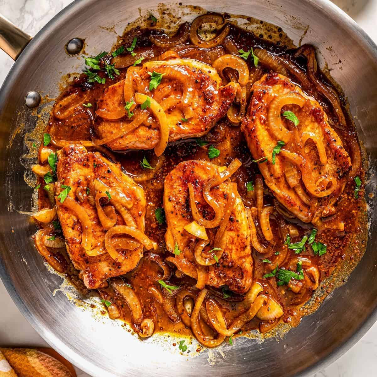 A large skillet with four cooked pork chops topped with onions and a mustard sauce.