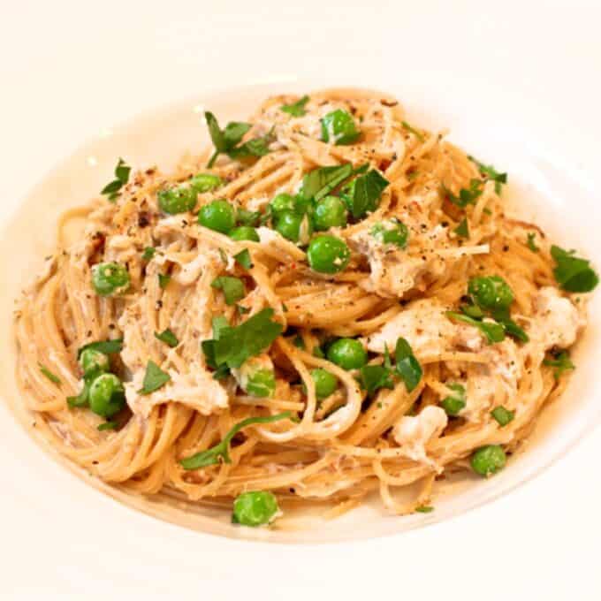 Close up of a low white bowl filled with a creamy crab pasta with green peas and sprinkled with chopped fresh parsley.