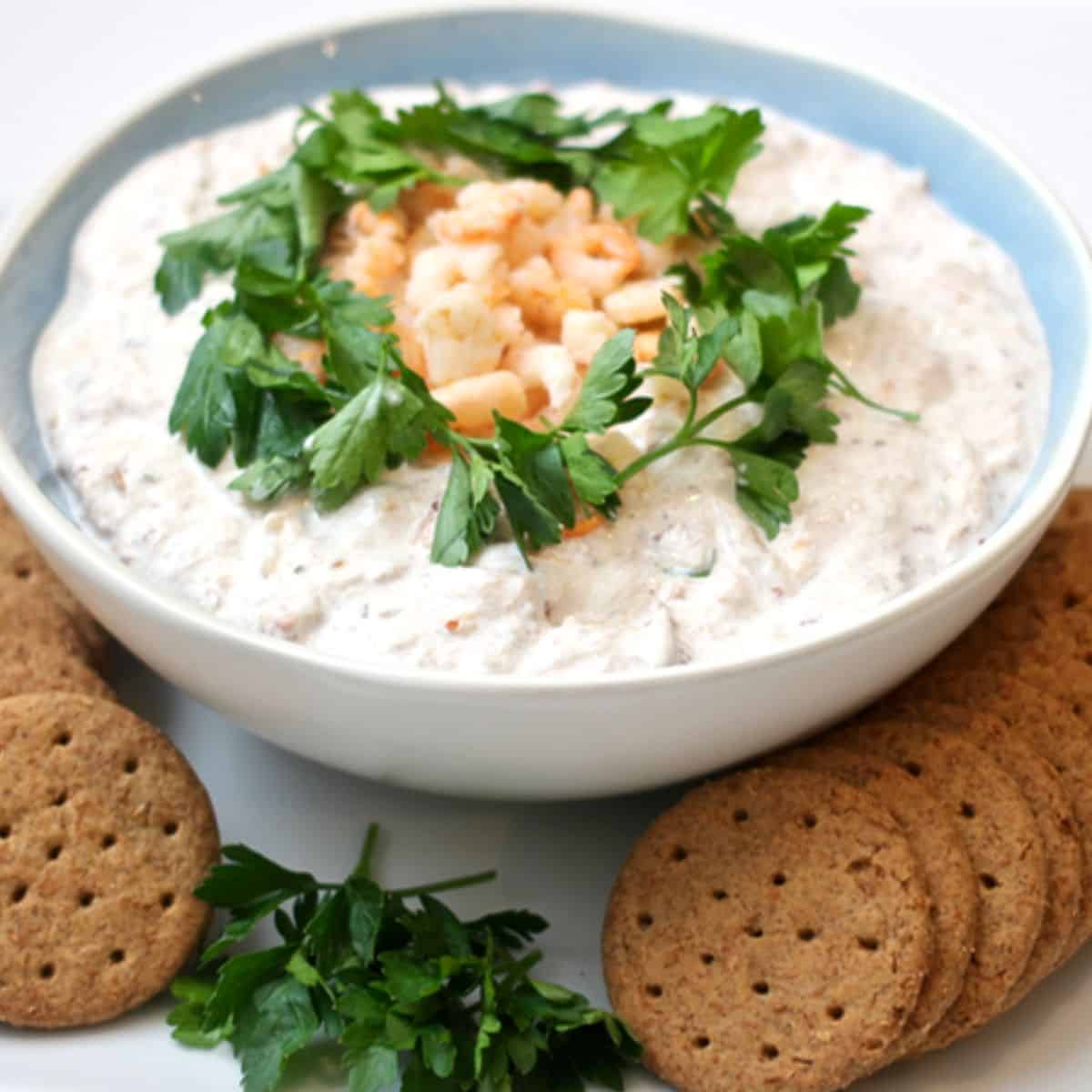 A blue and white bowl filled with a creamy crab dip appetizer topped with fresh parsley and chopped crab and crackers to the side for dipping.