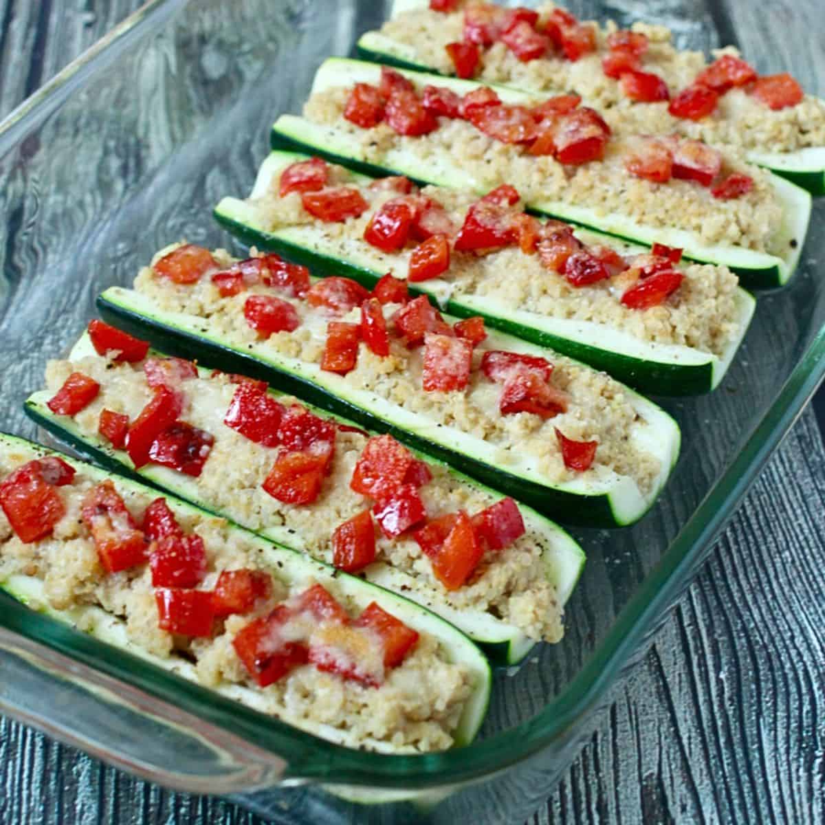 A glass baking dish with quinoa and hummus stuffed zucchini boats topped with roasted red peppers and Parmesan cheese.