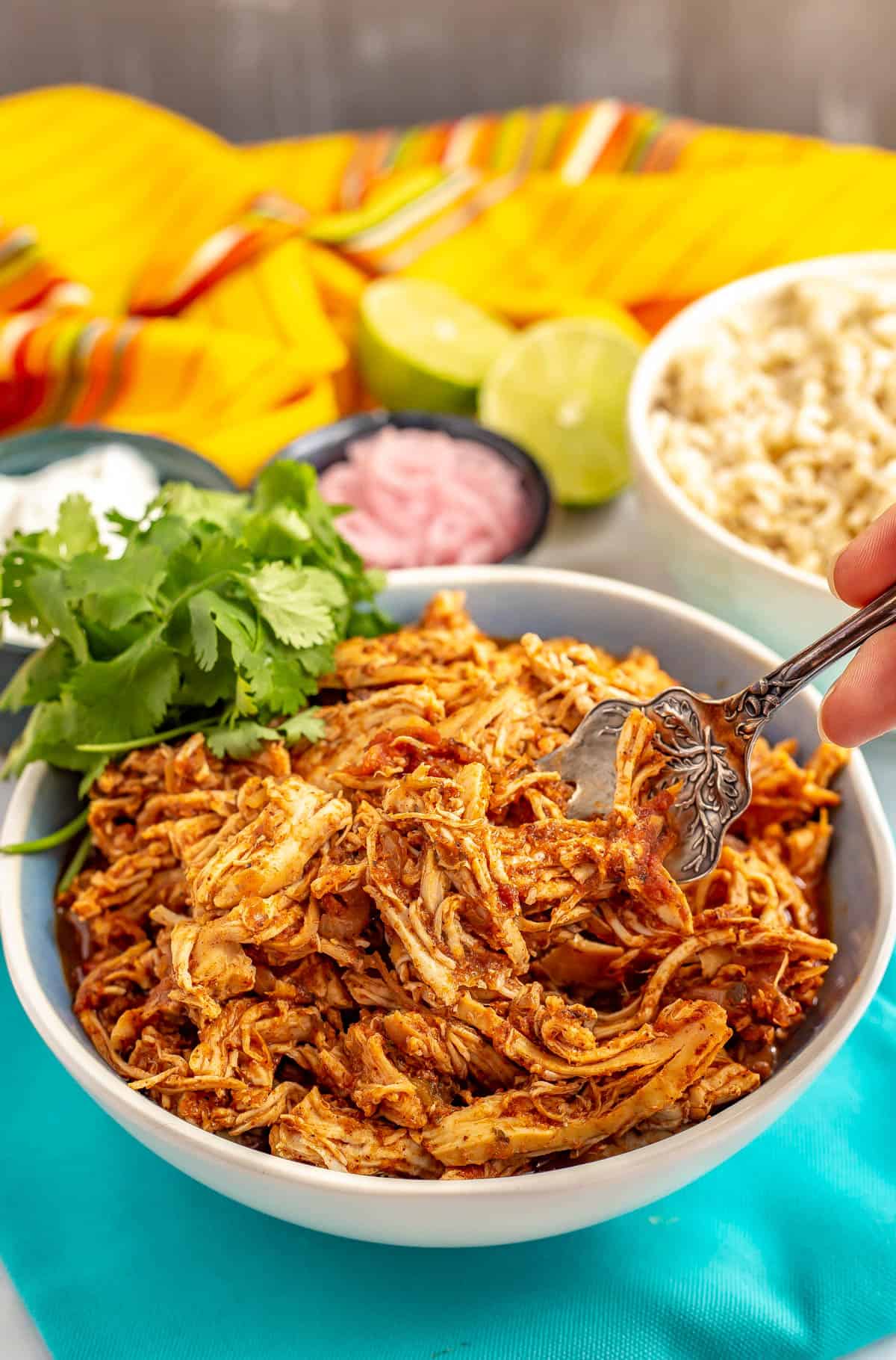 A serving fork scooping up some cooked shredded Mexican chicken from a bowl.