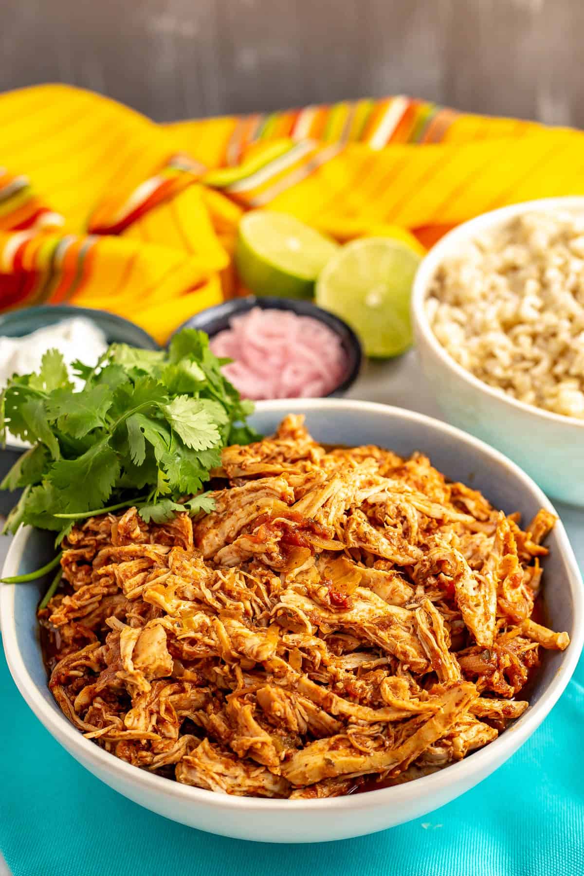 A blue bowl of slow cooker Mexican shredded chicken with cilantro for a garnish and bowls of brown rice, pickled red onions and sour cream in the background.
