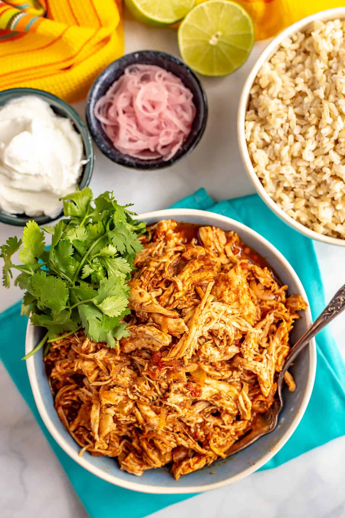 A fork resting in a bowl of crock pot Mexican shredded chicken alongside bowls of rice, sour cream and pickled red onions.