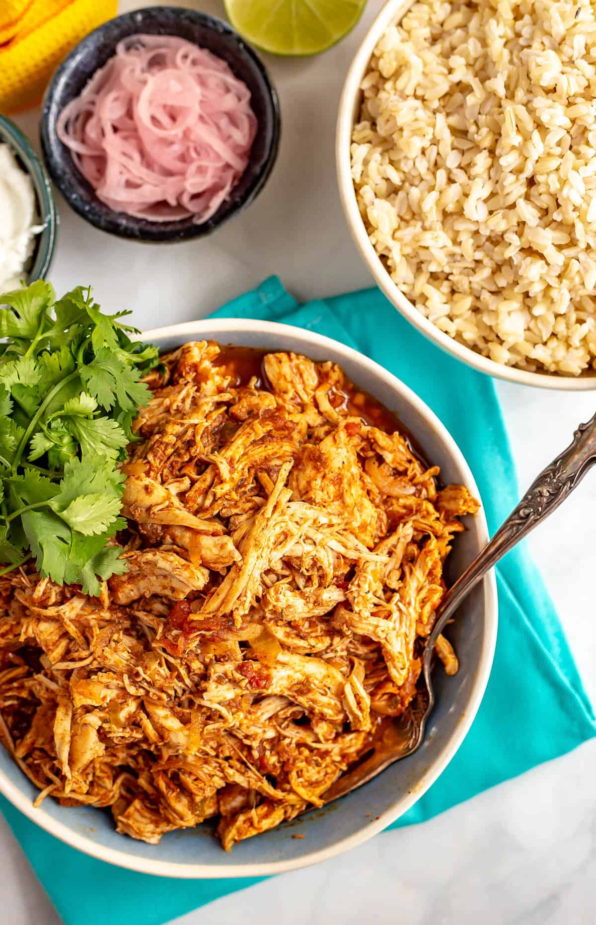 Close up of a fork resting in a bowl of crock pot Mexican shredded chicken alongside bowls of rice, sour cream and pickled red onions.