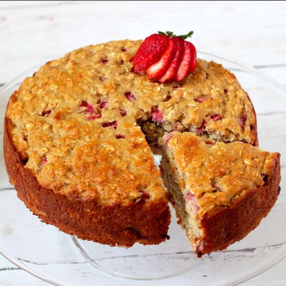 A tall strawberry cake on a glass cake stand with a large piece pulled out to one side.