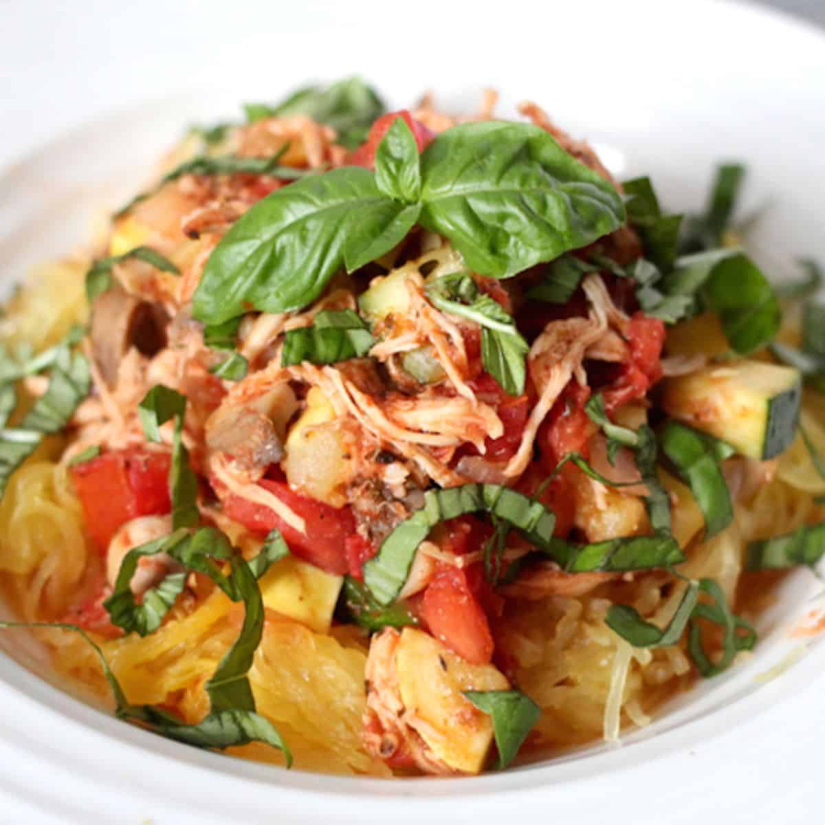 A low white bowl filled with spaghetti squash topped with a shredded chicken, tomato and veggie mixture with fresh basil on top.