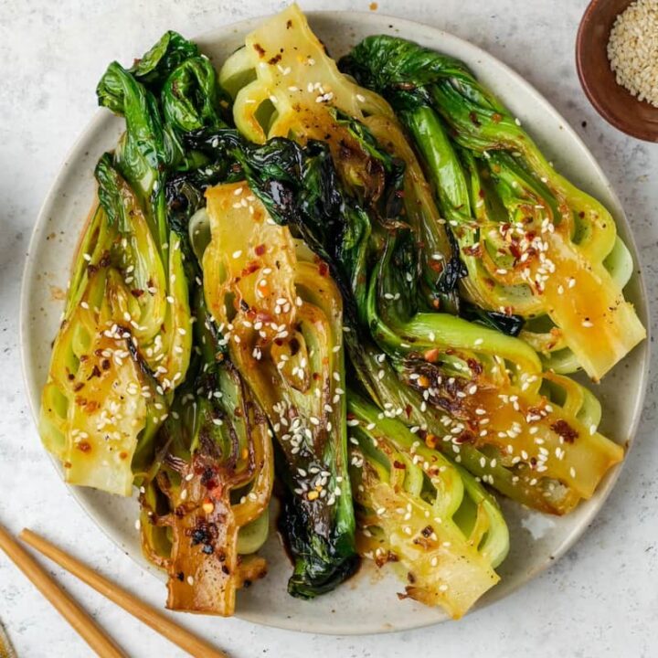 A round serving plate with baby bok choy in a soy sauce with soy sauce and sesame seeds in bowls to the side.