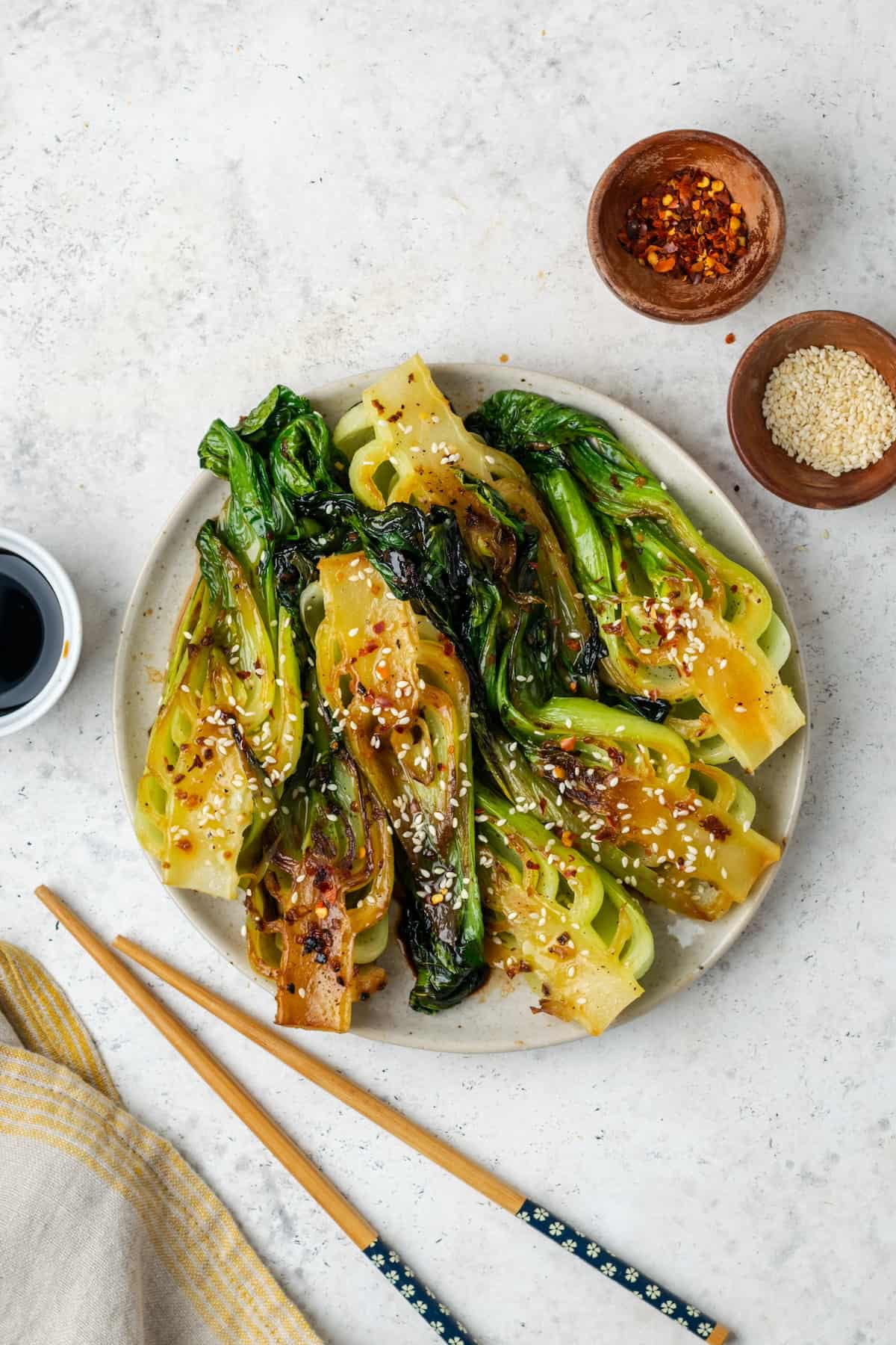 Baby bok choy with garlic and ginger served on a round platter with soy sauce, red pepper flakes and sesame seeds in bowls to the side.